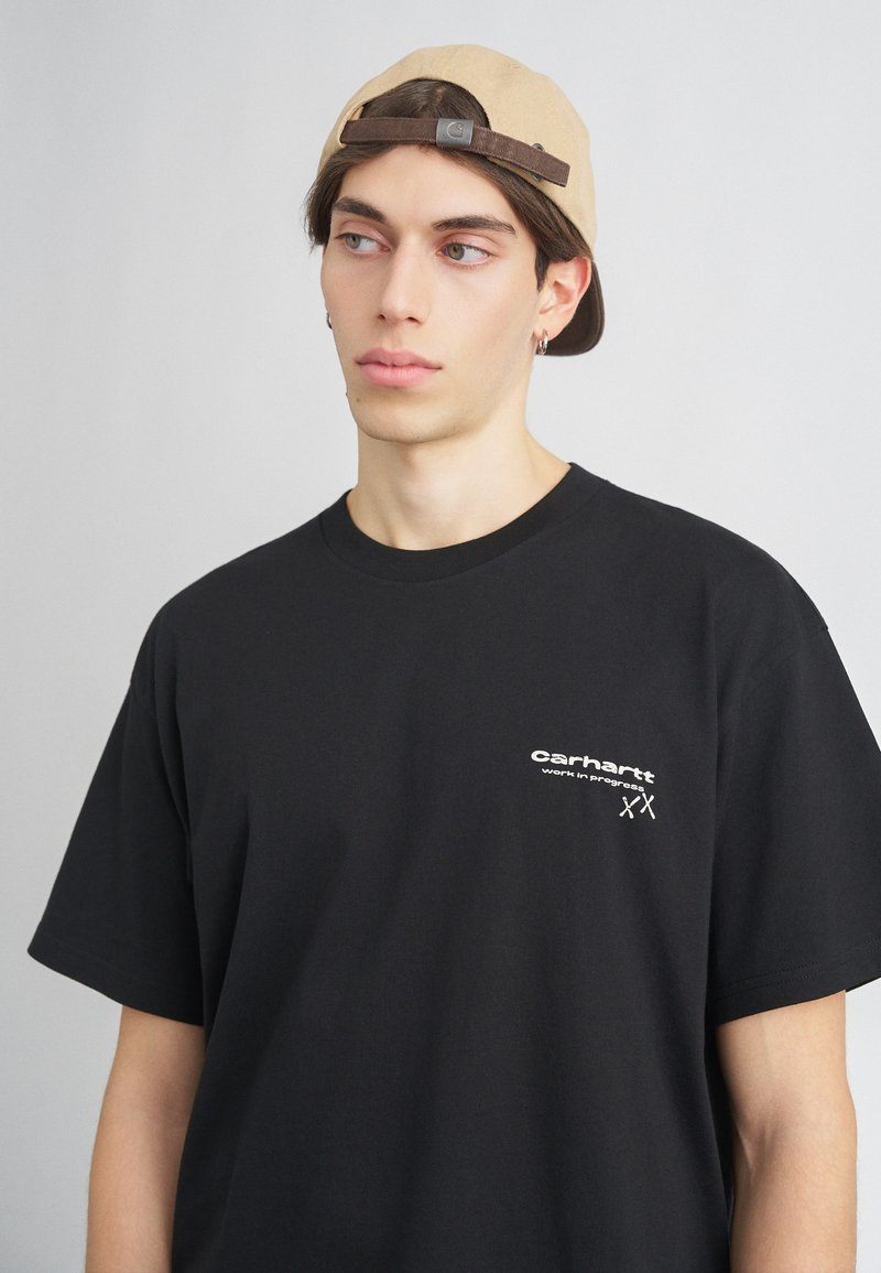 Camiseta de algodón negra con cuello redondo, mangas cortas y un logo blanco en el pecho. El modelo lleva una gorra de un tono beige claro.