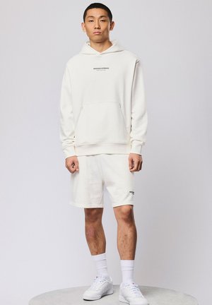 Junger Mann steht, trägt einen cremeweißen Hoodie und Shorts mit kleinem schwarzem Text, dazu weiße Socken und weiße Turnschuhe, neutraler Hintergrund.