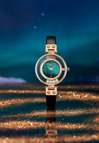 Reloj redondo con una esfera color aguamarina, hardware en tono dorado, correa de cuero negro y cinco cristales decorativos en la carátula.