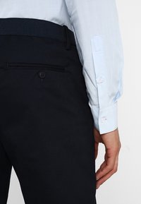 Chemise bleu clair à manches longues, associée à un pantalon bleu marine foncé avec une poche et des passants de ceinture, texture de tissu lisse.