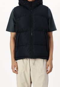 Czarna pikowana kamizelka z kapturem, z zamkiem błyskawicznym z przodu i dwiema bocznymi kieszeniami, noszona na ciemnozielonej koszulce z krótkim rękawem.