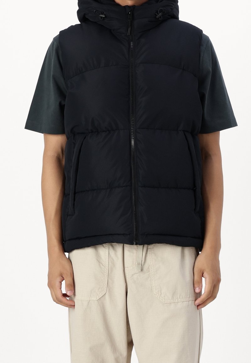 Czarna pikowana kamizelka z kapturem, z zamkiem błyskawicznym z przodu i dwiema bocznymi kieszeniami, noszona na ciemnozielonej koszulce z krótkim rękawem.