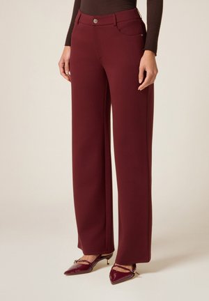 Femme portant un pantalon large taille haute bordeaux, associé à des talons pointus assortis et un haut à manches longues marron foncé.