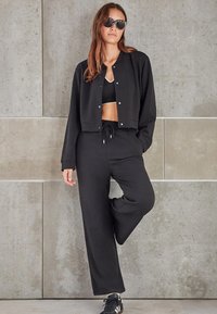 Schwarze cropped Jacke mit Knopfverschluss, kombiniert mit einer passenden schwarzen Jogginghose und schwarzen Sportschuhen. Sonnenbrillen vervollständigen den Look.