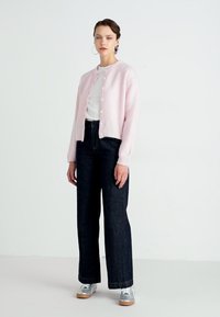 Απαλό ροζ, μαλακό κάρντιγκ με στρογγυλή λαιμόκοψη και κουμπί, συνδυασμένο με σκούρα φαρδιά τζιν και ασημένια αθλητικά παπούτσια. Απλές χρυσές σκουλαρίκια.