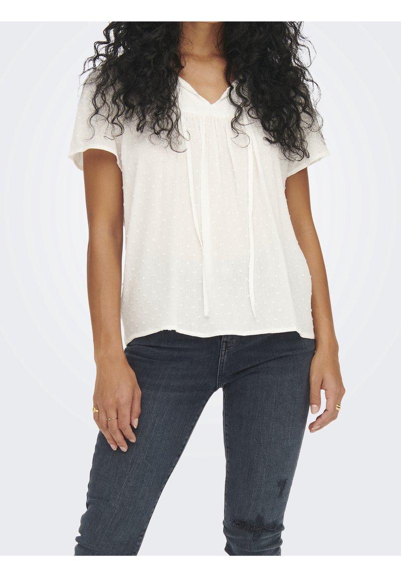 Valkoinen lyhythihainen pusero, jossa on teksturoidut pilkkupisteet ja V-pääntie. Edessä solmittava yksityiskohta ja väljä istuvuus. Yhdistetty tummiin denimfarkkuihin.
