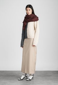 Saia longa de bege claro, suéter de malha creme com mangas cinzas, cachecol vermelho escuro, saltos strappy em preto e branco, textura suave ao longo de toda a peça.