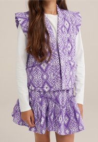 Lila mönstrad väst med volangärmar, matchad med en kjol med fransar i nederkant, tillverkad av lätt och texturerad tyg.