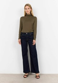 Olivgröns formsydda polotröja i kombination med blå vida jeans. Har en knappstängning, fickor och kontrasterande sömmar. Bärs med öppna klackar.
