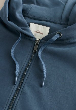Zils džemperis ar rāvējslēdzēju un kapuci, mīkstas tekstūras, ar auklu kapucē, sudraba rāvējslēdzēju un iekšpusē esošu etiķeti ar uzrakstu "NEXT SIGNATURE".