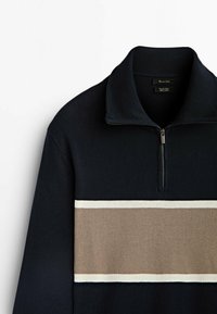 Pull-over bleu marine avec une bande horizontale beige et blanche sur la poitrine, col côtelé et fermeture éclair à mi-hauteur.