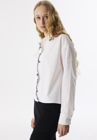 Camisa blanca de botones con detalles de ribete negro y un corte relajado, con mangas largas y dobladillo redondeado. Combinada con pantalones negros.
