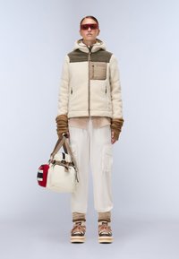 Chaqueta de forro polar en tono beige y crema con capucha y bolsillo frontal, combinada con pantalones cargo beige y zapatos de plataforma con estampado. Los accesorios incluyen un bolso.