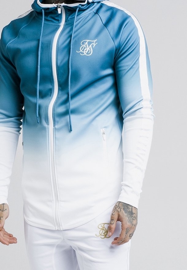 Hættetrøje i gradient blå og hvide farver med lynlås foran, sidelommer og et logo på brystet. Glat, tætsiddende stof.