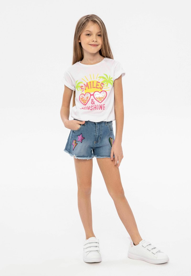 White t-shirt with colorful graphic featuring sunglasses and text, paired with denim shorts displaying various patch designs; white sneakers.