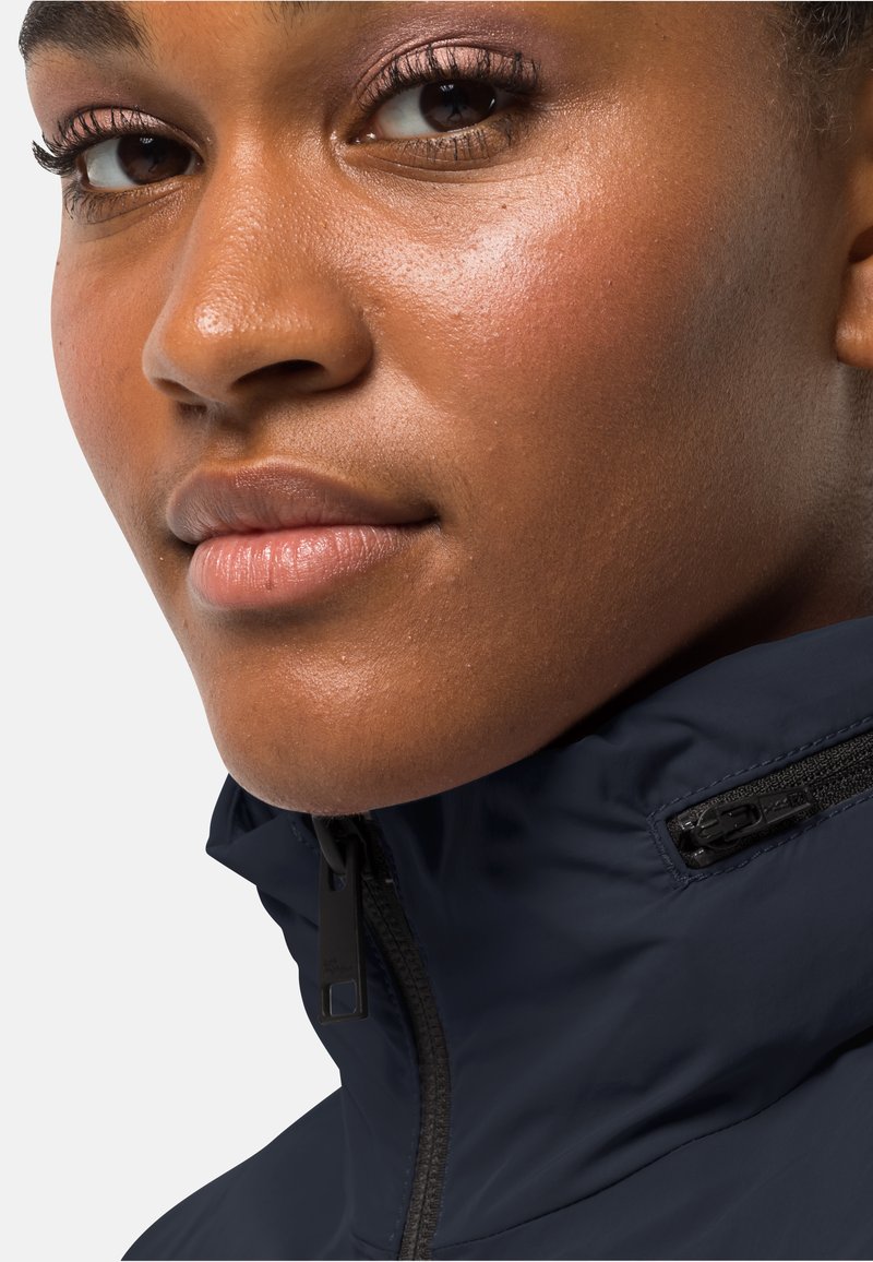 Marineblaue Jacke mit hochgeschlossenem Kragen, ausgestattet mit einem schwarzen Reißverschluss und strukturiertem Stoff; Nahaufnahme, die auf das Gesicht und die Details des Kragens fokussiert.