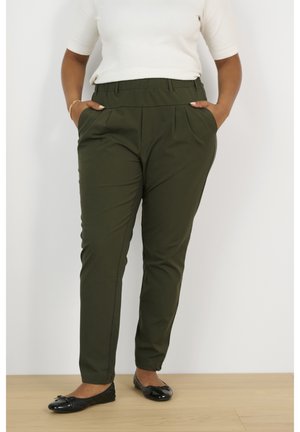 Vrouw draagt donker groene, op maat gemaakte broek met zakken en zwarte balletschoenen, staand op een houten vloer.