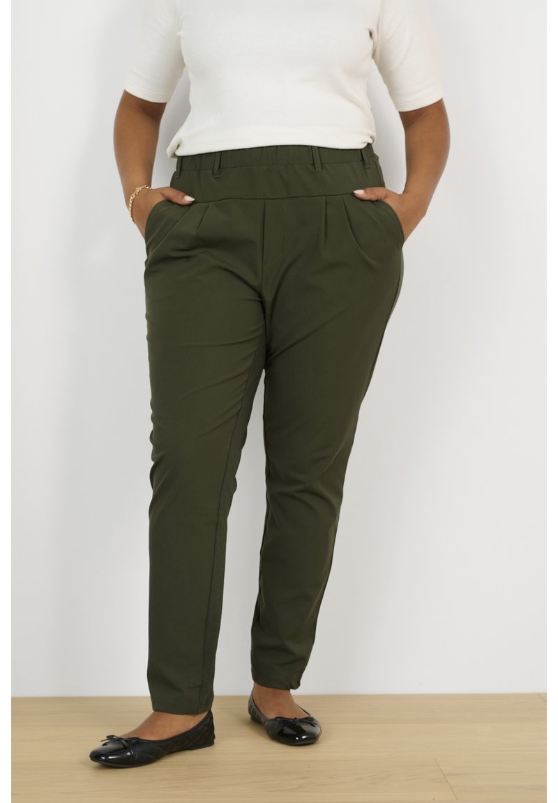 Femme portant un pantalon vert foncé sur mesure avec des poches et des ballerines noires, debout sur un sol en bois.