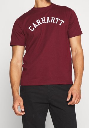 Mężczyzna ubrany w bordową koszulkę z krótkim rękawem z białym tekstem "CARHARTT" i czarne spodnie na jasnym tle.