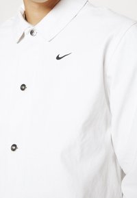 Närbild på en person som bär en vit skjorta med knappar och krage samt en liten svart Nike swoosh-logotyp på bröstet.