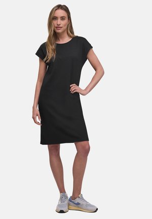 Frau mit langen blonden Haaren, die ein schwarzes knielanges Kleid mit kurzen Ärmeln und graue Turnschuhe trägt, steht mit einer Hand in der Hüfte.