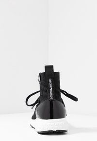 Sneaker nera con tomaia in tessuto a coste e tacco in vernice lucida. Presenta lacci rotondi e un logo sul retro. Suola bianca testurizzata.