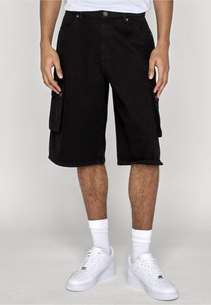 Mand iført sorte cargo-shorts til knæet, hvide crew-sokker og hvide sneakers, stående mod en ensartet lys baggrund.