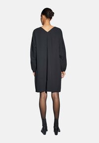 Zwarte jurk met lange mouwen, een lage V-rug, losse pasvorm en subtiele plooien. Gecombineerd met zwarte enkellaarsjes en transparante panty's.