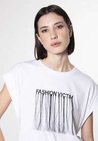 Hvid bomulds t-shirt med korte ærmer, der har sort "FASHION VICTIM" tekst og frynse design. Glat tekstur, afslappet pasform.