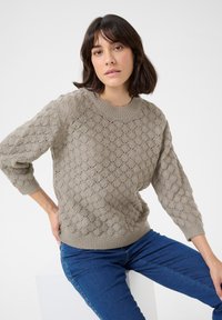 Hellbrauner, gestrickter Pullover mit strukturiertem Muster, geripptem Halsausschnitt und Bündchen, kombiniert mit blauen Jeans. Modell sitzt vor einem schlichten Hintergrund.