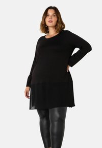 Musta pitkähiha paita kerroksellisella suunnittelulla, jossa on läpikuultava alaosa ja pyöreä pääntie, yhdistettynä kiiltäviin mustiin leggings-housuihin.