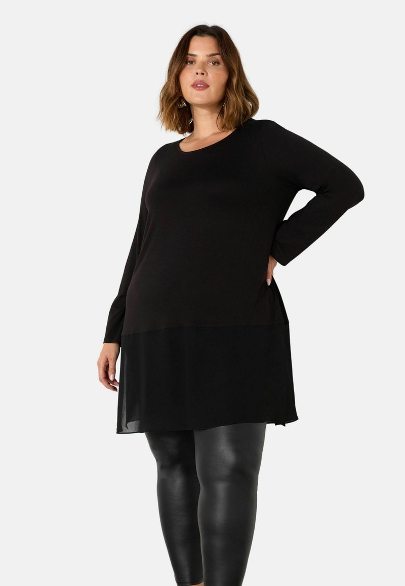 Musta pitkähiha paita kerroksellisella suunnittelulla, jossa on läpikuultava alaosa ja pyöreä pääntie, yhdistettynä kiiltäviin mustiin leggings-housuihin.