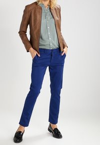 Bruine leren jas, lichtgroen overhemd met knopen, blauwe broek, zwarte loafers. Focus op texturen en kleuren.