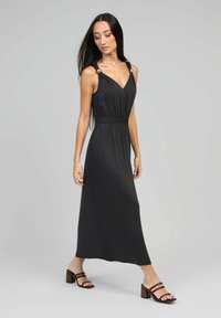 Femme marchant dans une robe maxi noire sans manches avec des détails de boutons dorés aux épaules, portant des sandales à talons bruns sur un fond gris.