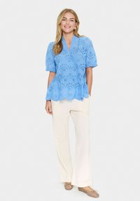 Blusa peplum azzurro chiaro con ritagli floreali, maniche a palloncino corte e orlo scallop, abbinata a pantaloni cream a gamba larga e ballerine marroni.