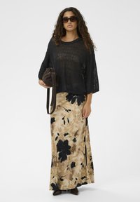 Sieviete, kura valkā tumšus saulesbrilles un melnu caurspīdīgu topu ar bēšas krāsas ziedu maxi svārkiem, turot tumši brūnu rokassomu pret baltu fona.