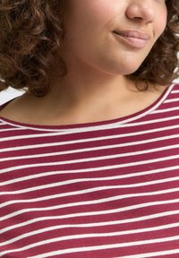 Vrouw met krullend bruin haar die een bordeauxrood en wit gestreept shirt draagt, zacht glimlachend met gesloten lippen, nek en schouder zichtbaar.