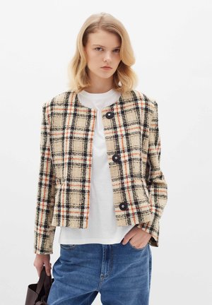 Veste à carreaux beige avec des accents noirs et orange, col rond, quatre boutons, tissu texturé et deux poches latérales. Portée par-dessus une chemise blanche.