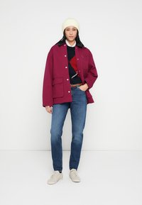 Mørkerød jakke med trykknapper, sidelommer; brukt over en marineblå genser med rødt diamantmønster. Kombinert med blå jeans og hvite joggesko.