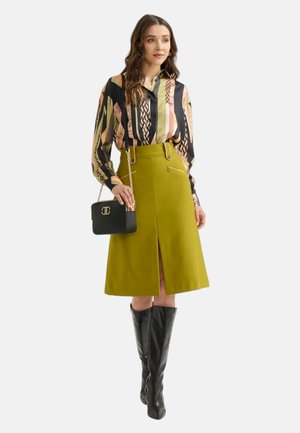 Blusa scura a fantasia con maniche lunghe, gonna giallo-verde fino al ginocchio con spacco frontale, stivali neri fino al ginocchio e borsa a spalla nera.