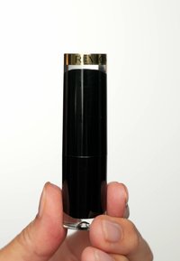 Botón de lápiz labial cilíndrico negro con tapa dorada brillante. La base clara revela el lápiz labial. El nombre de la marca "Revlon" está grabado en la tapa. Textura suave.