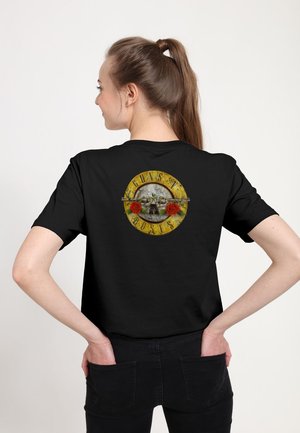 Kobieta z brązowym kucykiem nosi czarną koszulkę z vintage logo Guns N' Roses przedstawiającym dwie czerwone róże i skrzyżowane pistolety na plecach.