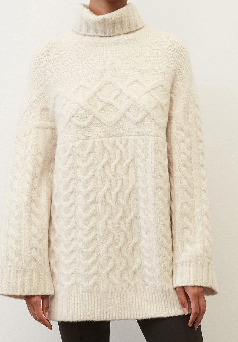 Pull à col roulé en maille torsadée couleur crème, à manches longues et poignets côtelés, porté par une personne portant un pantalon foncé.