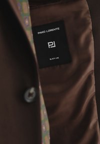 Wnętrze brązowej kurtki z czarną metką "PAKO LORENTE" i "BLACK LINE", obok wzorzystej podszewki w zielonych i niebieskich odcieniach.