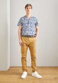 Cămașă cu mânecă scurtă, cu un model albastru și alb, pantaloni chinos bej și adidași albi. Cămașa are un design geometric, iar pantaloni au o textură netedă.