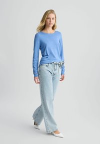Hellblaues langärmliges Oberteil mit Rundhalsausschnitt und dezentem Muster. Kombiniert mit hoch taillierten, locker sitzenden, hellen Jeans und weißen High Heels.