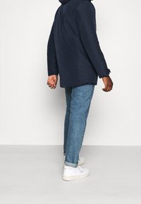 Mand iført en marineblå jakke, lyseblå opsmøgede jeans og hvide sneakers, der står foran en ensfarvet hvid baggrund.