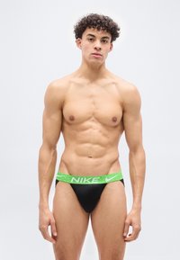 Νεαρός άνδρας με σγουρά μαλλιά στέκεται γυμνόστηθος φορώντας μαύρα αθλητικά εσώρουχα με πράσινη ζώνη Nike, μπροστά σε ένα απλό λευκό φόντο.