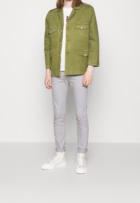 Chaqueta verde oliva con dos bolsillos en el pecho, combinada con pantalones ajustados gris claro y zapatillas altas blancas. Textura de tela suave.