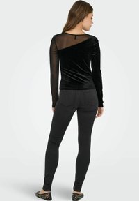 Žena stojící zády, oblečená v černém sametovém topu s průhlednými rukávy a černých skinny džínách, na jednobarevném bílém pozadí.